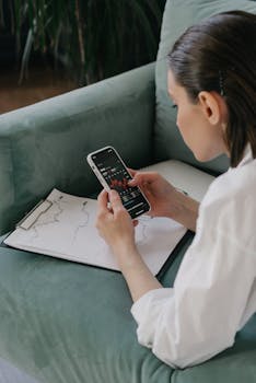 A woman examines financial stock data on her smartphone, sitting indoors with a graph on paper.