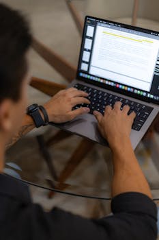 Adult man working on a laptop at a glass table, focused on typing.